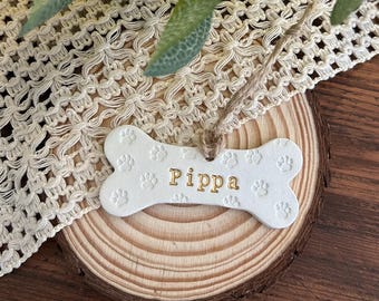 May include: A white bone-shaped ornament featuring embossed paw prints across the surface and the name Pippa printed in gold lettering. The ornament includes a brown twine loop attached to the top for hanging purposes.