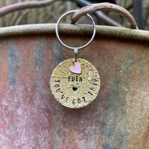 May include: Gold-toned keychain with a round tag that reads "You've Got This" with a small heart in the centre. The tag also has the name "Eden" and a pink heart charm.