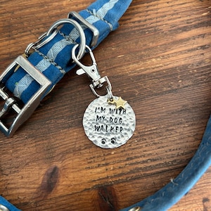 May include: A blue fabric dog collar with a silver-toned metal buckle and a circular, hammered-texture metal tag attached by a lobster clasp. The tag features the stamped text I'M WITH MY DOG WALKER, a small gold-toned star, and two black paw prints.