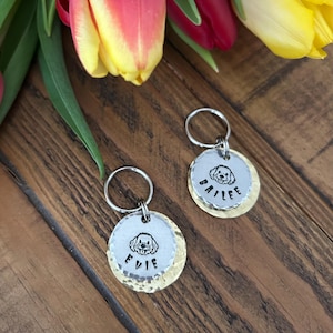 Puede incluir: Dos llaveros personalizados con chapas para perros. Cada llavero tiene un disco superior plateado y un disco inferior martillado de color dorado. Las etiquetas presentan una ilustración de perro y un nombre. Los llaveros están sobre una superficie de madera con flores coloridas en el fondo.