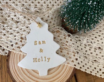Könnte beinhalten: Ein weißer, baumförmiger Keramikschmuck mit dem goldgeprägten Text "Sam & Holly" und einem kleinen goldenen Sternanhänger an der Spitze. Der Schmuck hängt an einer dünnen Schnur und ist auf einer runden Holzscheibe platziert.