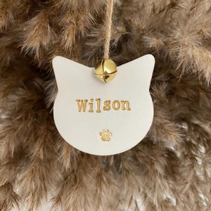 Op de afbeelding: Een wit katvormig ornament met de naam "Wilson" in gouden letters en een gouden pootafdruk. Een klein gouden belletje is aan de bovenkant bevestigd met een touwtje. Het ornament hangt tegen een achtergrond van gedroogde planten.