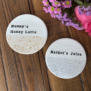 Peut inclure: Deux dessous de verre circulaires avec une moitié supérieure blanche texturée et une moitié inférieure mouchetée. L'un porte le texte noir Mummy's Honey Latte, l'autre Margot's Juice, tous deux dans une police de style machine à écrire.