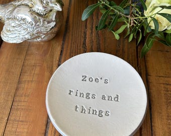 May include: A round white ceramic trinket dish featuring the stamped text Zoe's rings and things in the center. The dish is positioned near a metallic silver bird figurine and green foliage, showcasing a clean and simple design for storage.