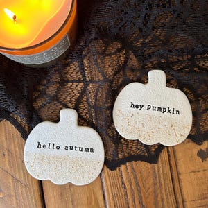 Könnte beinhalten: Zwei cremeweiße, kürbisförmige Untersetzer mit einer texturierten unteren Hälfte. Ein Untersetzer trägt den schwarzen Stempelaufdruck "hello autumn", der andere den Aufdruck "hey Pumpkin" in einer einfachen Schreibmaschinenschrift.
