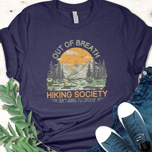 May include: A navy blue t-shirt with a vintage-style graphic of a mountain landscape. The graphic says "Out of Breath Hiking Society Don't Worry, I'll Catch Up".