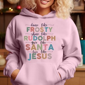 Puede incluir: Sudadera con capucha rosa claro con bolsillo delantero y el texto "dance like FROSTY, shine like RUDOLPH, give like SANTA, love like JESUS" en varios colores. La sudadera es de estilo jersey.