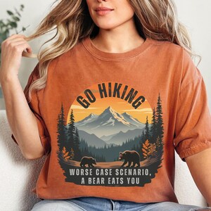 May include: A rust-colored t-shirt with a graphic of a mountain range, trees, and two bears. The text "Go Hiking" is in a circle above the mountain range. The text "Worse case scenario, a bear eats you" is below the graphic.