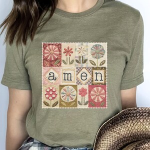 May include: Heather olive green t-shirt with a floral patchwork design. The design features various flowers and the word "amen" in a square pattern. The shirt is made of a soft material.