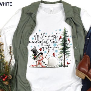 Puede incluir: Camiseta blanca de manga larga con un diseño de muñeco de nieve, sombrero de copa, bufanda y el texto "It's the most wonderful time of the year!". El diseño incluye un árbol de Navidad, cardenales y copos de nieve.