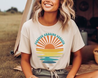 May include: A woman wearing a beige t-shirt with a retro sunset graphic and the text "FOREVER CHASING SUNSETS". She is sitting outdoors with a white mug in her hand.