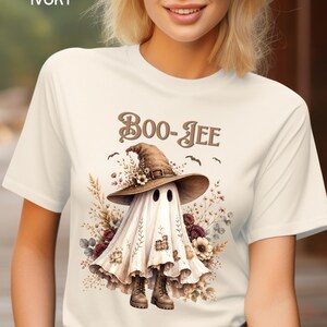 May include: An ivory t-shirt featuring a graphic of a ghost wearing a brown witch hat and boots, surrounded by dried flowers and small bats. The text "BOO-JEE" is printed in brown above the ghost, which has patches on its white sheet body.