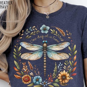 May include: A navy blue t-shirt with a floral design featuring a dragonfly and the text "One day at a time".