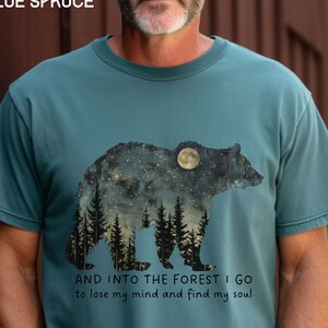 Pu&ograve; includere: Una t-shirt blu turchese con una grafica di un orso in silhouette con un cielo notturno stellato e una foresta al suo interno. Il testo "AND INTO THE FOREST I GO to lose my mind and find my soul" &egrave; stampato sotto la grafica.