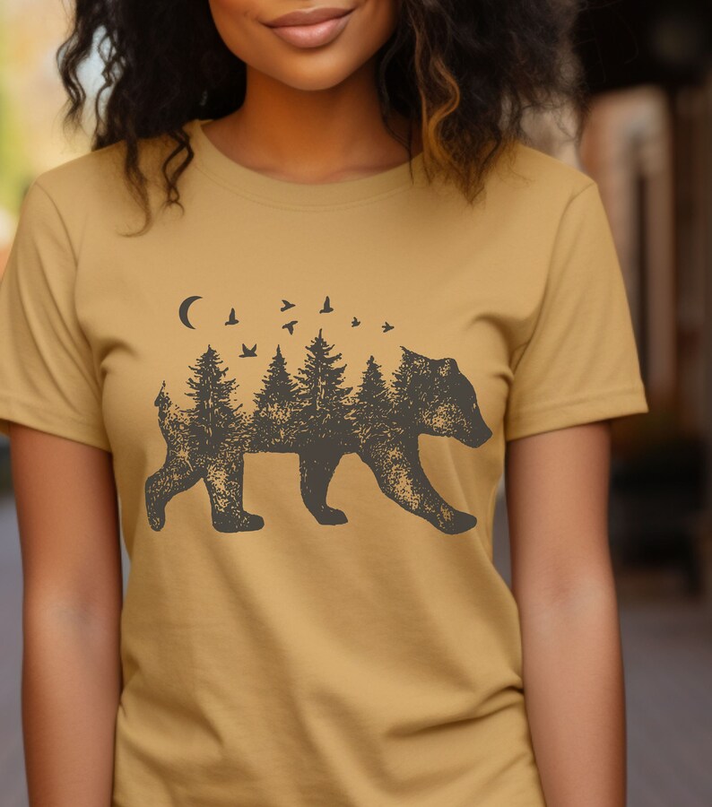 May include: A mustard yellow t-shirt with a black silhouette of a bear. The bear is made up of trees and a crescent moon. There are small birds flying above the trees.