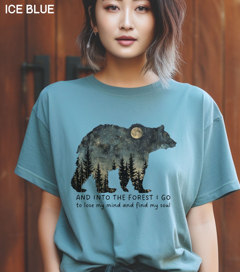 Pu&ograve; includere: Una maglietta blu chiaro con una silhouette nera di un orso in piedi in un bosco con una luna piena dietro. Il testo "AND INTO THE FOREST I GO to lose my mind and find my soul" &egrave; stampato sotto l'orso.
