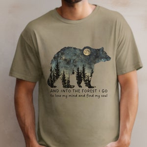 Pu&ograve; includere: T-shirt color kaki con la silhouette di un orso con una luna e stelle al suo interno. Il testo "AND INTO THE FOREST I GO to lose my mind and find my soul" &egrave; stampato sotto l'orso.