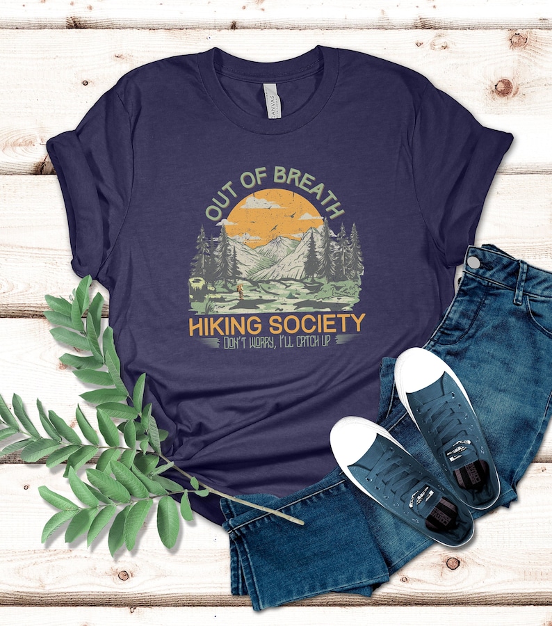 May include: A navy blue t-shirt with a vintage-style graphic of a mountain landscape. The graphic says "Out of Breath Hiking Society Don't Worry, I'll Catch Up".
