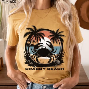 May include: A mustard yellow t-shirt with a graphic of a crab on a beach with palm trees and a sunset. The text "Crabby Beach" is printed below the graphic.