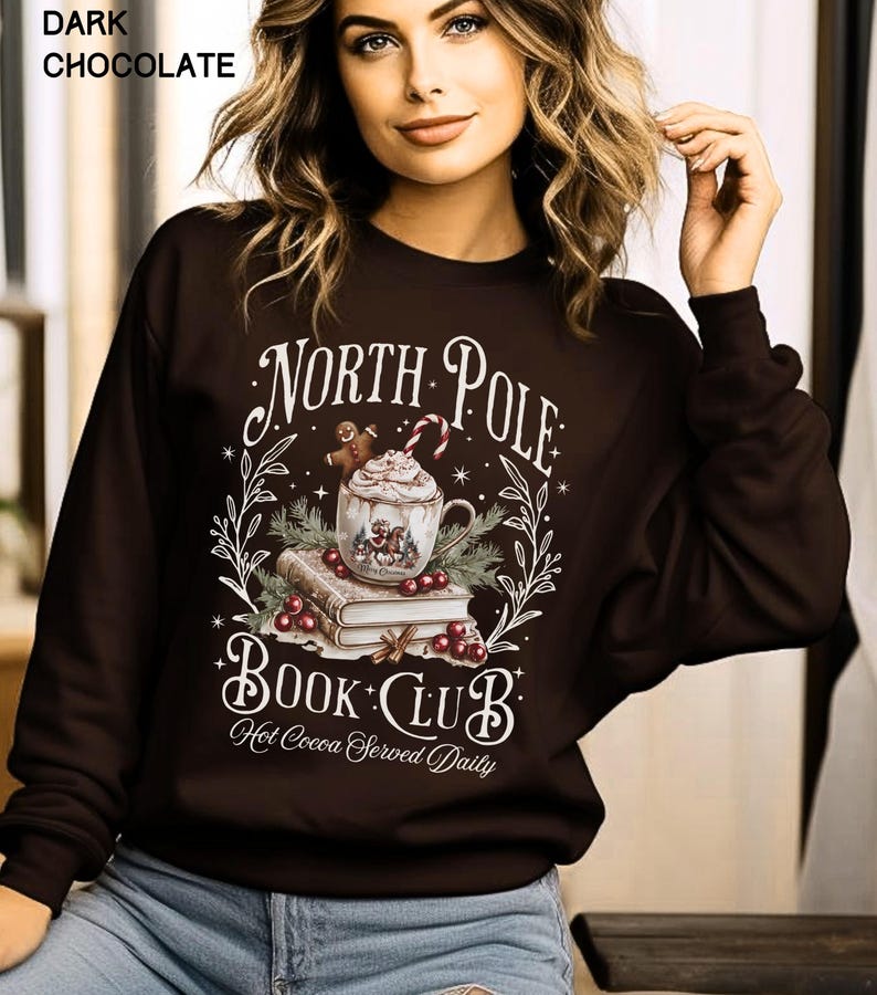 Puede incluir: Sudadera de color chocolate oscuro con el texto "North Pole Book Club" y un gr&aacute;fico de una taza de chocolate caliente, libros y bayas. Las palabras "Hot Cocoa Served Daily" tambi&eacute;n est&aacute;n impresas en la sudadera.