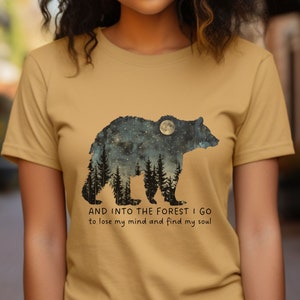 Pu&ograve; includere: Una maglietta color senape con la silhouette di un orso con un cielo stellato notturno e una luna piena all'interno dell'orso. Il testo "AND INTO THE FOREST I GO to lose my mind and find my soul" &egrave; stampato sotto l'orso.