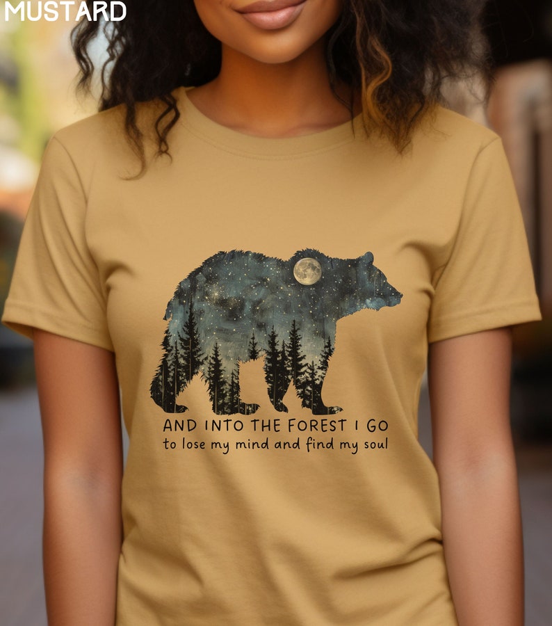 Pu&ograve; includere: Una maglietta color senape con la silhouette di un orso con un cielo stellato notturno e una luna piena all'interno dell'orso. Il testo "AND INTO THE FOREST I GO to lose my mind and find my soul" &egrave; stampato sotto l'orso.