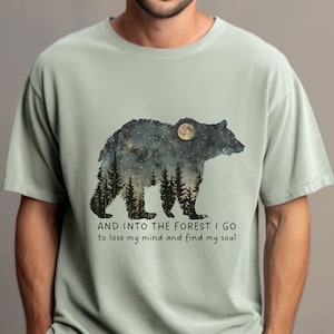 Pu&ograve; includere: Una maglietta verde chiaro con una grafica di un orso in silhouette con un cielo notturno stellato e una luna piena all'interno dell'orso. Il testo "AND INTO THE FOREST I GO to lose my mind and find my soul" &egrave; stampato sotto l'orso.