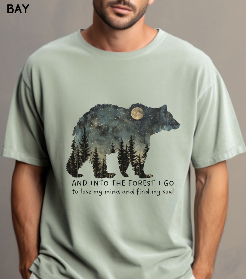 Pu&ograve; includere: Una maglietta verde chiaro con una grafica di un orso in silhouette con un cielo notturno stellato e una luna piena all'interno dell'orso. Il testo "AND INTO THE FOREST I GO to lose my mind and find my soul" &egrave; stampato sotto l'orso.
