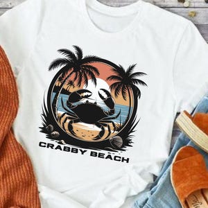 May include: White t-shirt with a graphic of a crab on a beach with palm trees and the text "Crabby Beach".