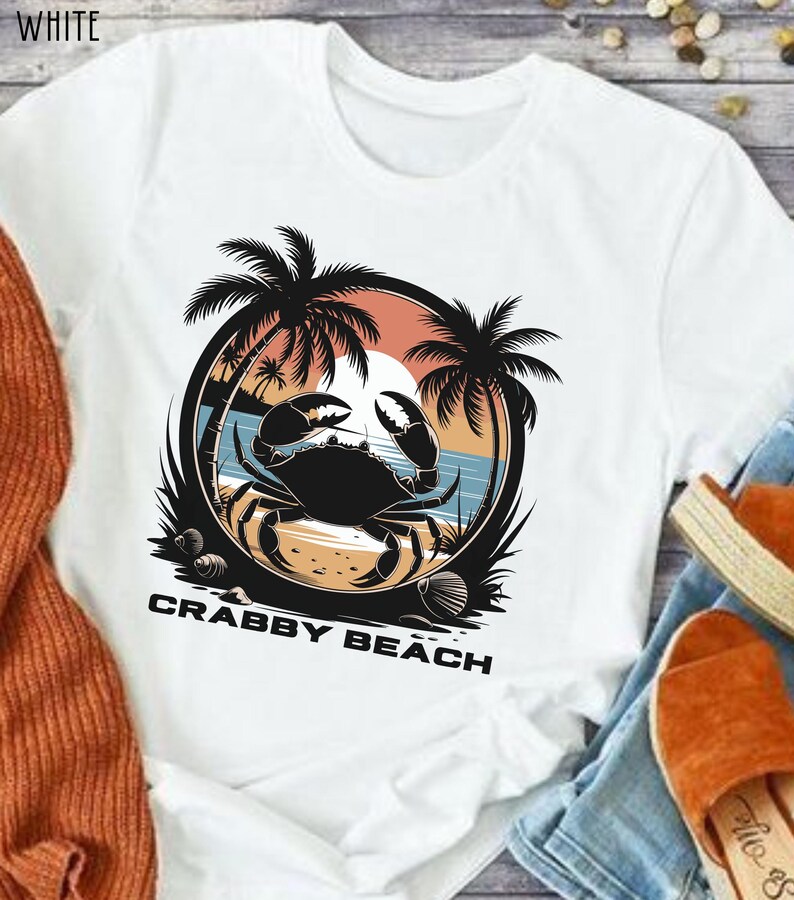 May include: White t-shirt with a graphic of a crab on a beach with palm trees and the text "Crabby Beach".