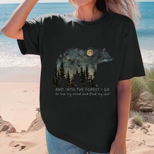 Pu&ograve; includere: Maglietta nera con una grafica di una silhouette di orso con un cielo notturno stellato e una luna piena all'interno. Il testo "AND INTO THE FOREST I GO to lose my mind and find my soul" &egrave; stampato sotto la grafica.