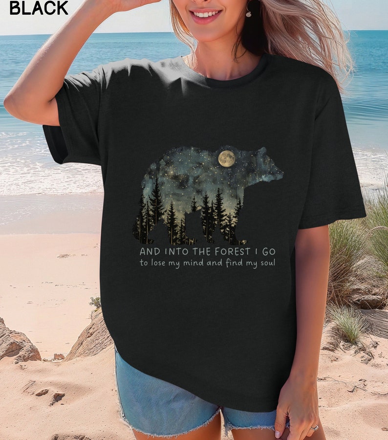 Pu&ograve; includere: Maglietta nera con una grafica di una silhouette di orso con un cielo notturno stellato e una luna piena all'interno. Il testo "AND INTO THE FOREST I GO to lose my mind and find my soul" &egrave; stampato sotto la grafica.