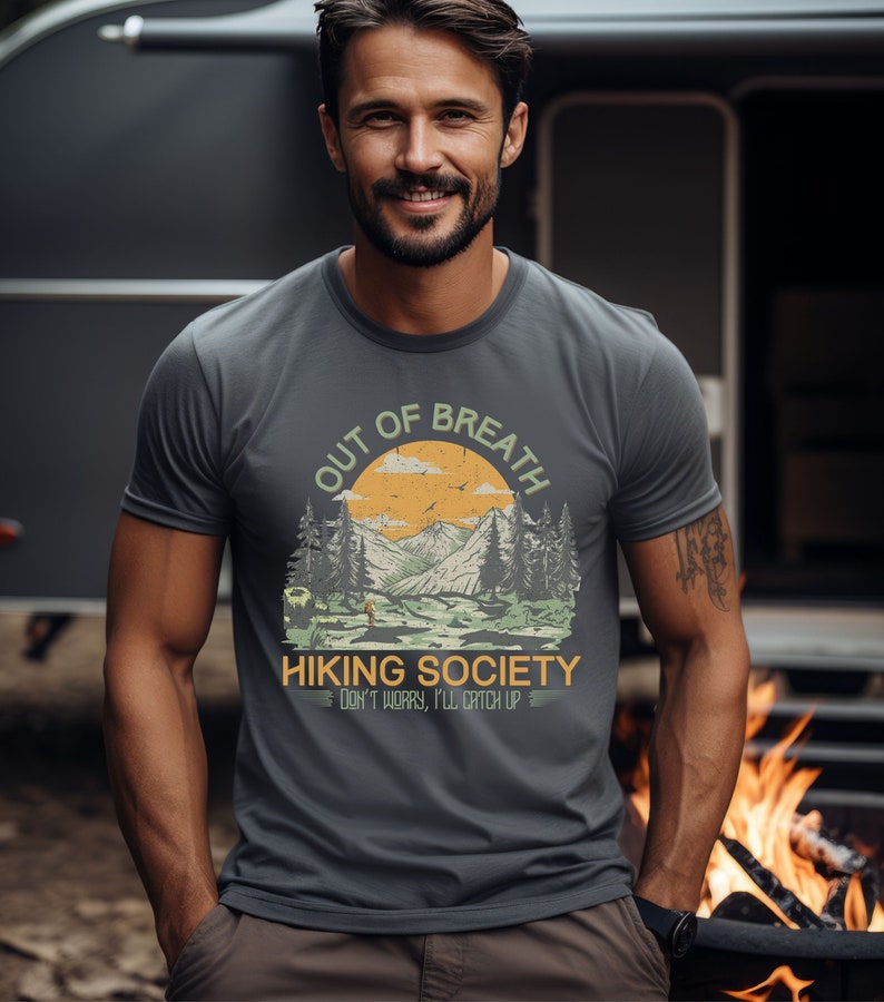May include: A dark gray t-shirt with a vintage-style graphic of a mountain landscape. The text "Out of Breath Hiking Society Don't Worry, I'll Catch Up" is printed on the shirt.