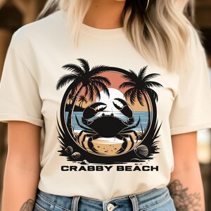 May include: A white t-shirt with a graphic of a crab on a beach with palm trees and a sunset. The text "Crabby Beach" is printed below the graphic.