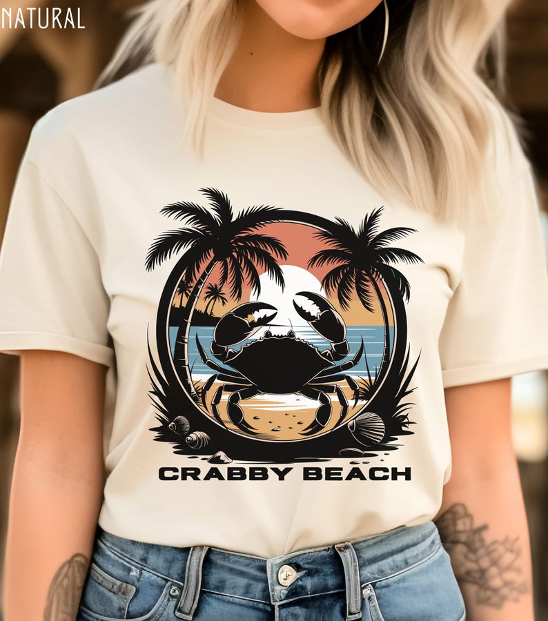 May include: A white t-shirt with a graphic of a crab on a beach with palm trees and a sunset. The text "Crabby Beach" is printed below the graphic.