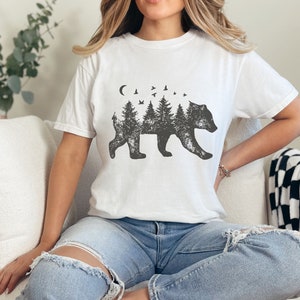 May include: White t-shirt with a black and white graphic of a bear silhouette with trees and birds inside the bear. The bear is walking to the right.
