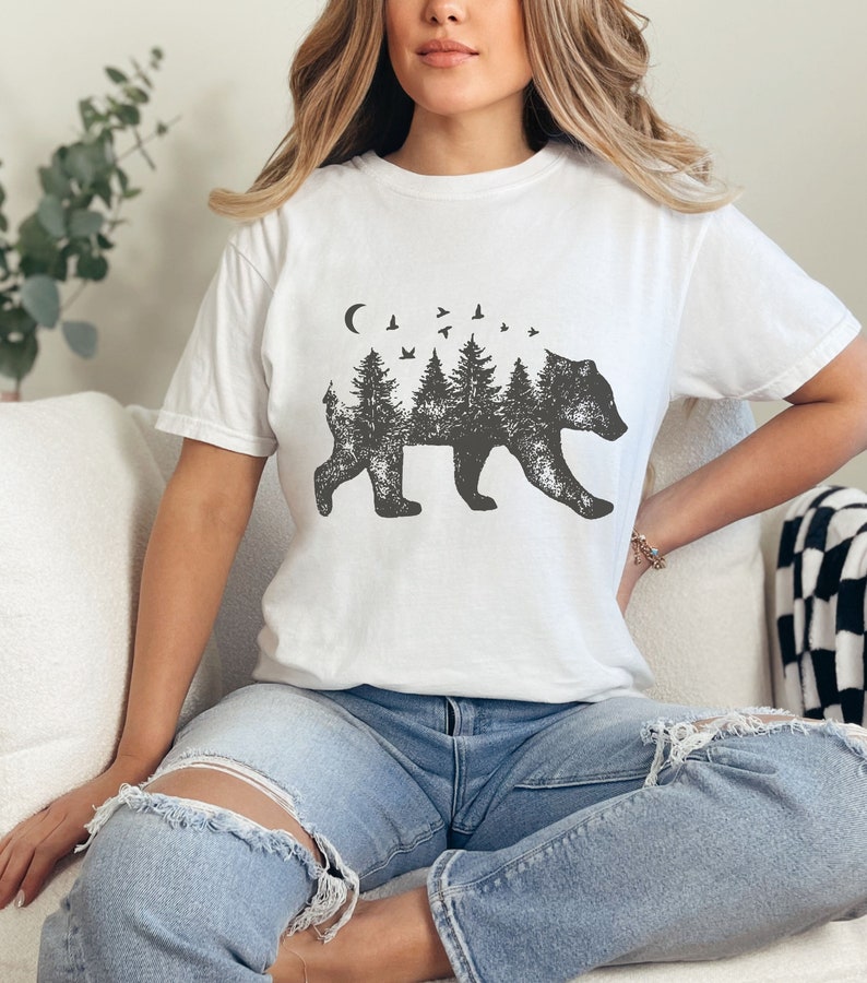 May include: White t-shirt with a black and white graphic of a bear silhouette with trees and birds inside the bear. The bear is walking to the right.