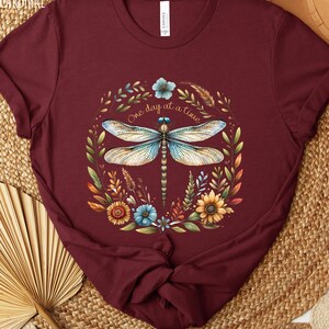 May include: A maroon t-shirt with a dragonfly and floral design. The dragonfly is blue and white with brown veins. The flowers are yellow, orange, blue, and green. The text "One day at a time" is written in a cursive font.