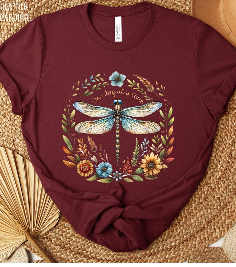 May include: A maroon t-shirt with a dragonfly and floral design. The dragonfly is blue and white with brown veins. The flowers are yellow, orange, blue, and green. The text "One day at a time" is written in a cursive font.