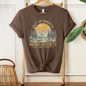 May include: Brown t-shirt with a graphic of a mountain landscape and the text "Out of Breath Hiking Society Don't Worry, I'll Catch Up".