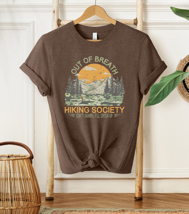 May include: Brown t-shirt with a graphic of a mountain landscape and the text "Out of Breath Hiking Society Don't Worry, I'll Catch Up".