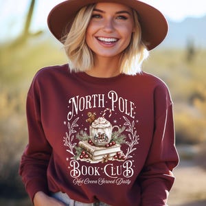 Puede incluir: Sudadera granate con un dise&ntilde;o que dice "North Pole Book Club" con un dise&ntilde;o de chocolate caliente. El dise&ntilde;o incluye una taza de chocolate caliente, libros y decoraciones festivas. La palabra "MAROON" est&aacute; impresa en la esquina superior izquierda.