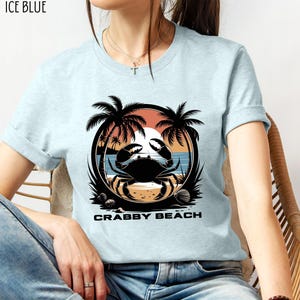 May include: A light blue t-shirt with a graphic of a crab on a beach with palm trees and the text "Crabby Beach".