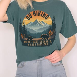 May include: A teal blue t-shirt with a graphic of a mountain range and two bears. The graphic is surrounded by a circle with the text "Go Hiking" at the top. The text "Worse case scenario, a bear eats you" is printed below the graphic.