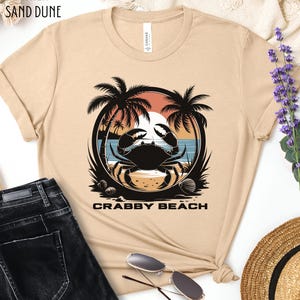 May include: A beige t-shirt with a graphic of a crab on a beach with palm trees and the text "Crabby Beach".