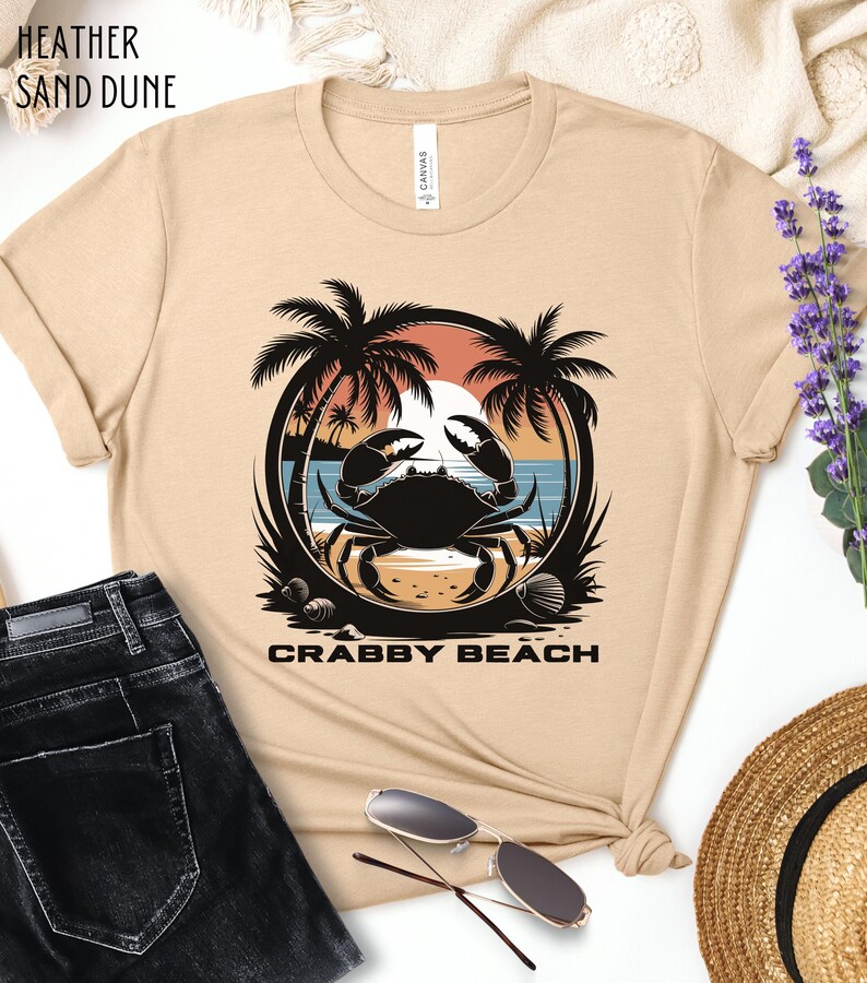 May include: A beige t-shirt with a graphic of a crab on a beach with palm trees and the text "Crabby Beach".