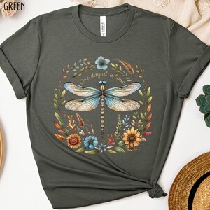 May include: A heather military green t-shirt with a dragonfly and floral design. The dragonfly is blue and brown with a gold outline. The flowers are yellow, orange, red, and blue. The text "One day at a time" is written in gold lettering.