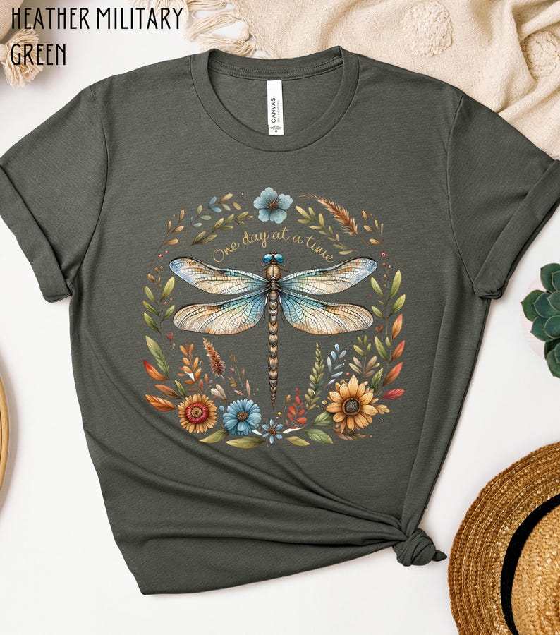 May include: A heather military green t-shirt with a dragonfly and floral design. The dragonfly is blue and brown with a gold outline. The flowers are yellow, orange, red, and blue. The text "One day at a time" is written in gold lettering.