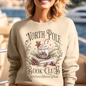 Puede incluir: Sudadera color arena con un gr&aacute;fico festivo. El dise&ntilde;o incluye las palabras "North Pole Book Club" con una taza de chocolate caliente, un hombre de jengibre y un bast&oacute;n de caramelo. Tambi&eacute;n est&aacute; presente la frase "Hot Cocoa Served Daily".