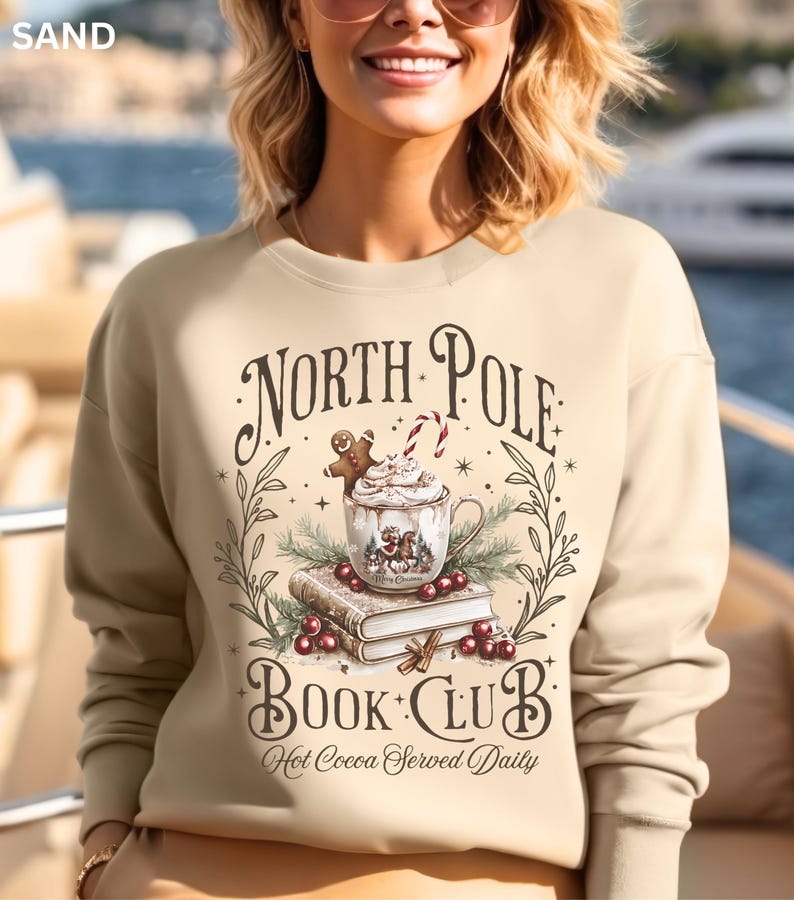 Puede incluir: Sudadera color arena con un gr&aacute;fico festivo. El dise&ntilde;o incluye las palabras "North Pole Book Club" con una taza de chocolate caliente, un hombre de jengibre y un bast&oacute;n de caramelo. Tambi&eacute;n est&aacute; presente la frase "Hot Cocoa Served Daily".